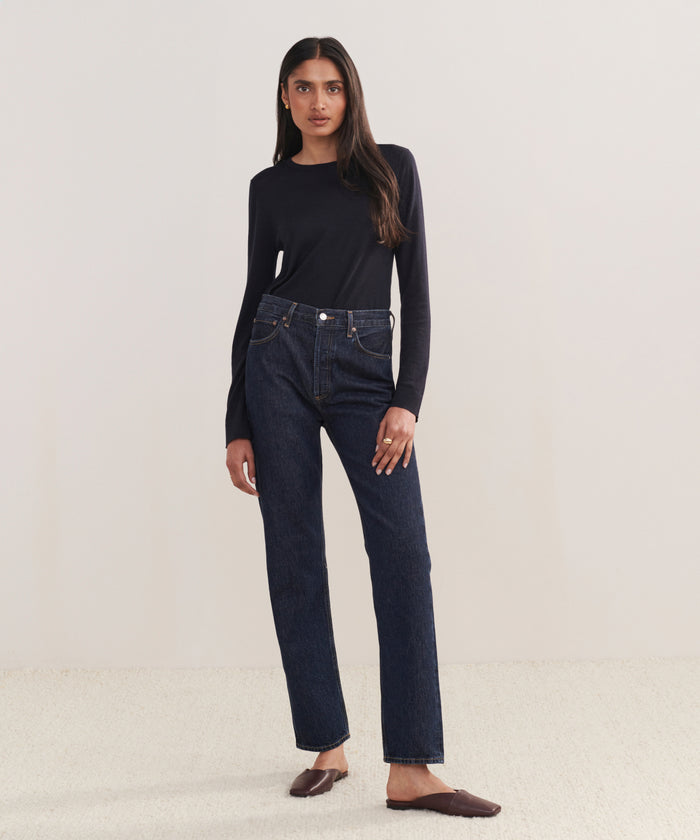 A woman with long straight hair poses confidently against a light background, wearing a fitted black long-sleeve top, 90s Pinch Waist Long Jean in dark blue, and brown flats.