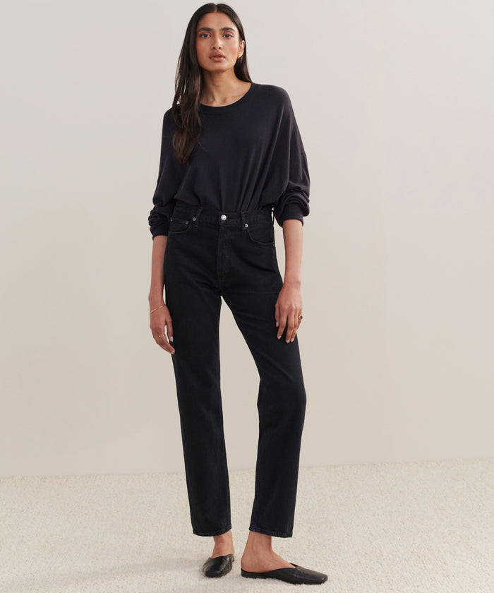 A woman with her hair down and a neutral expression stands against a plain background, wearing a loose black long-sleeve top tucked into 90s Pinch Waist Long Jeans and black slip-on shoes.