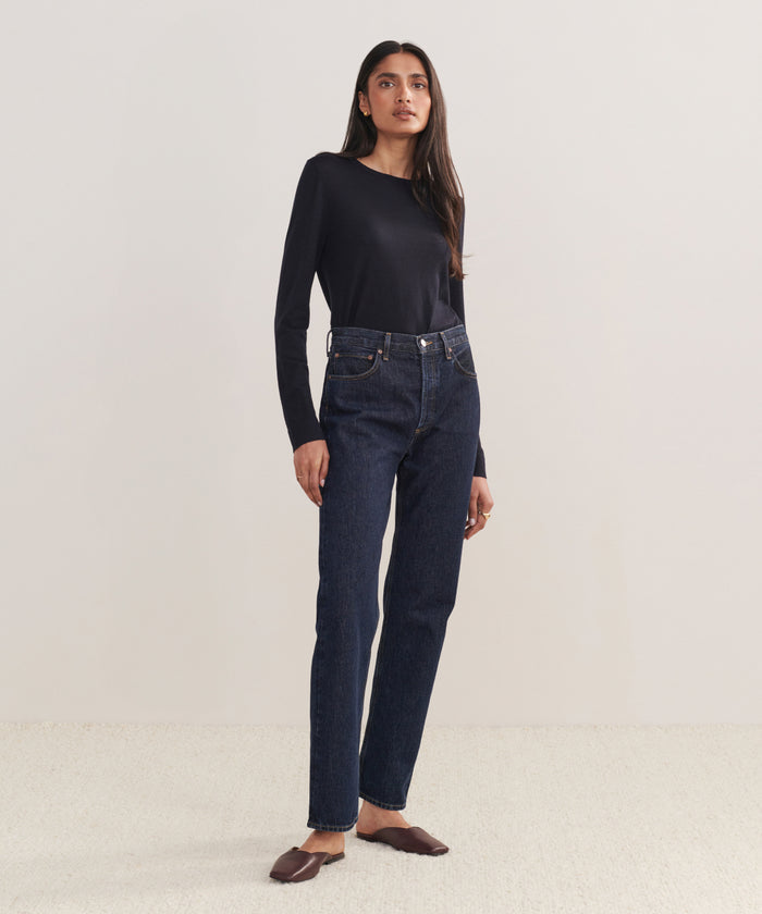 A woman with long, straight hair and a neutral expression stands against a plain background wearing a black long-sleeve top, brown flats, and the 90s Pinch Waist Long Jean in high-waisted dark blue organic cotton.