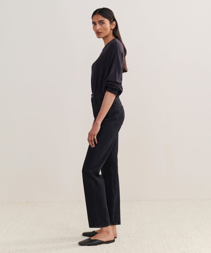 A woman with long, dark hair stands in profile against a light background, wearing a long-sleeve dark top, black flats, and the 90s Pinch Waist Long Jean. Her arms rest naturally at her sides.
