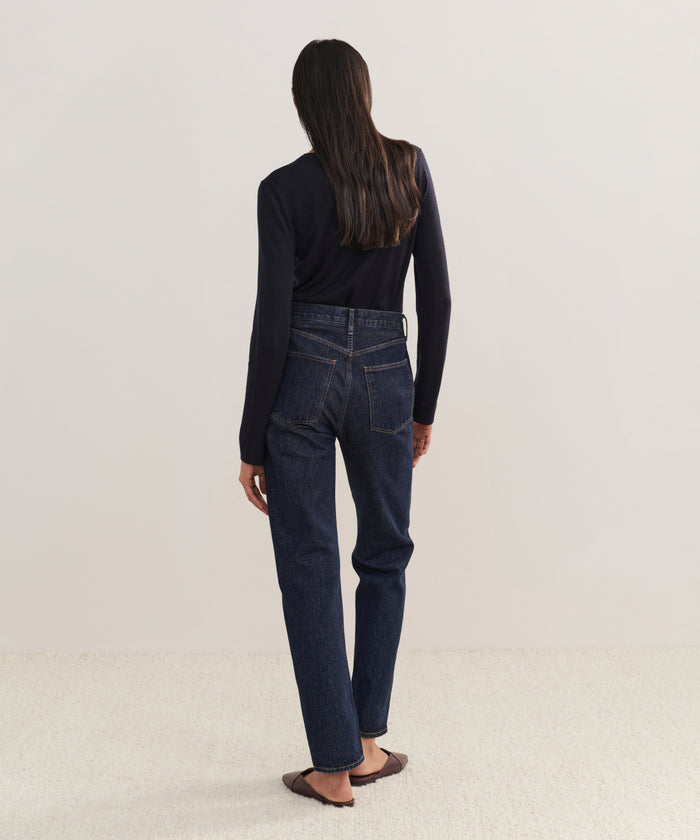 A person with long dark hair, in a black long-sleeve top, 90s Pinch Waist Long Jeans, and brown flats stands facing away from the camera against a plain light background.