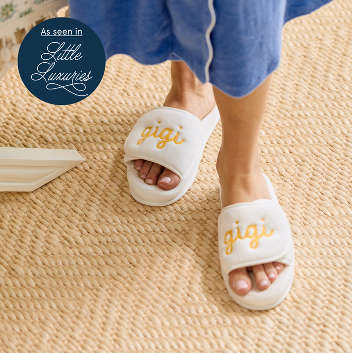 A person wearing white slide slippers with the word 'gigi' embroidered in yellow on the straps, standing on a woven beige rug. The person is also wearing blue pajama pants. There's a round blue tag that says, "As seen in Little Luxuries.'