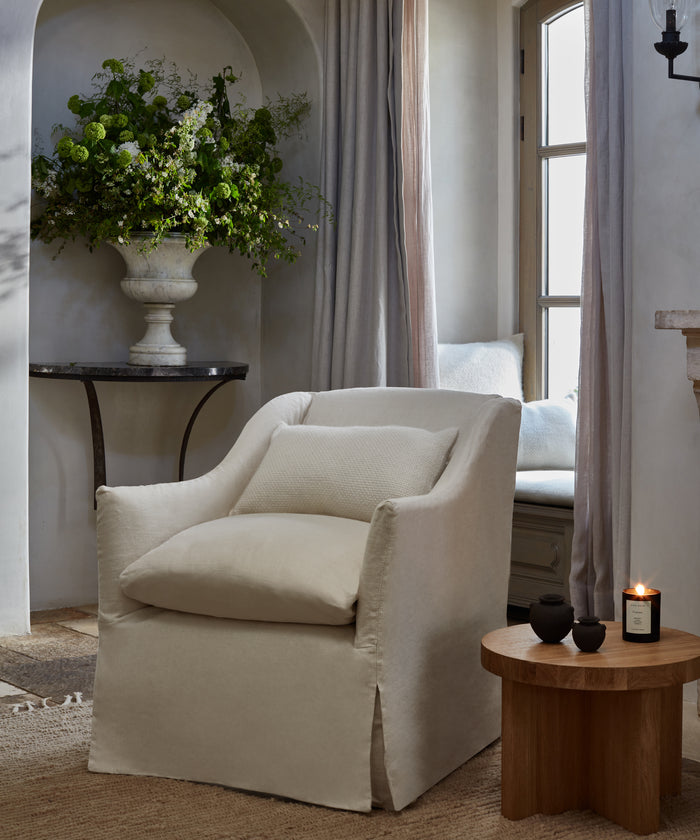 A cozy ivory armchair with the Alpaca Basketweave Lumbar Pillow sits beside a wooden side table holding a lit candle and small cups. Sheer curtains frame the window, while a lush green floral arrangement in a vase adds elegance.