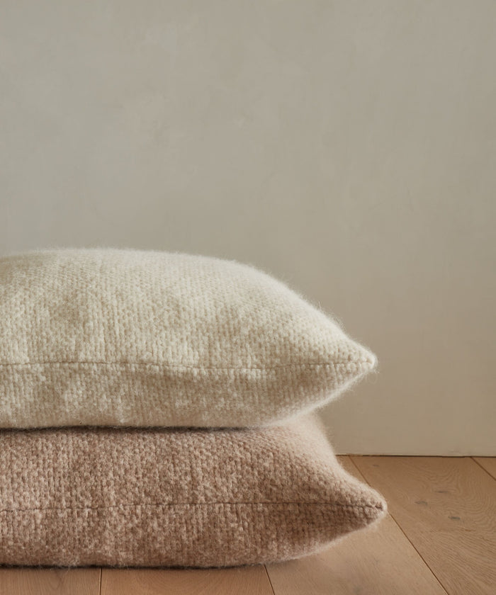 Two throw pillows stacked on a wooden floor against a light beige wall; the top is a cream-colored super soft pillow, while the bottom is an Alpaca Basketweave Pillow in cozy beige or dusty pink.