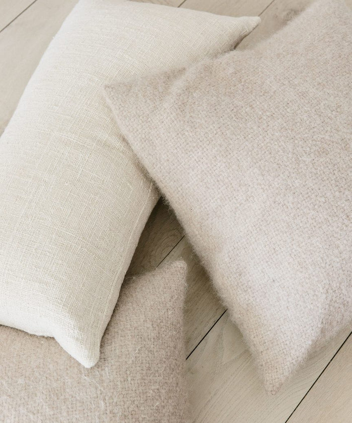 The Alpaca Basketweave Pillow, along with two neutral-toned pillows, sits on a light wooden floor. Their inviting textures and handmade in Peru quality make them perfect for minimalist or Scandinavian-inspired spaces.