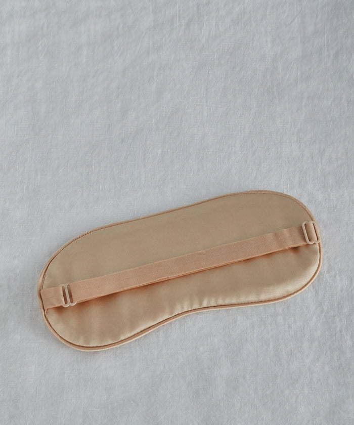 The Alpaca Eye Mask, made of beige silk with an elastic strap, rests on a light gray, textured alpaca wool surface.