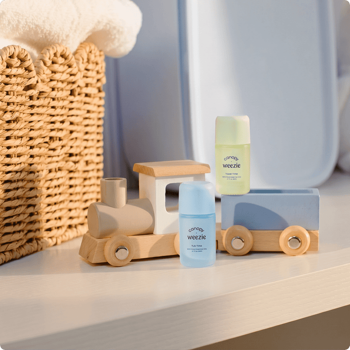 Three Weezie baby skincare bottles styled on a wooden toy train set next to a woven basket on a white shelf.