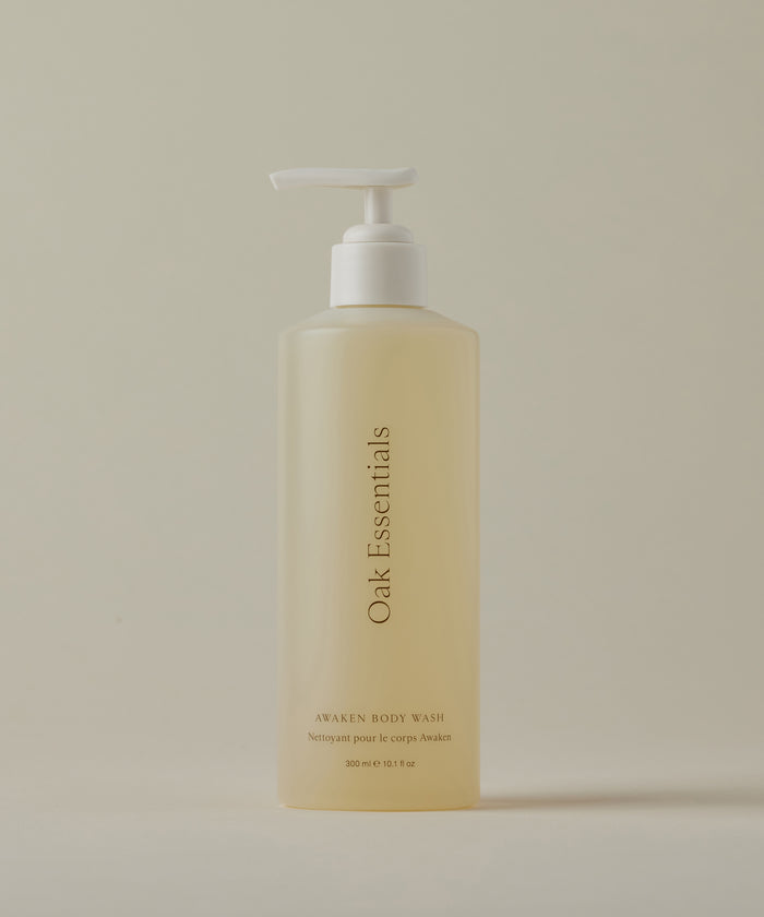 A translucent bottle with a white pump, labeled Awaken Body Wash, is displayed against a plain, light beige background.