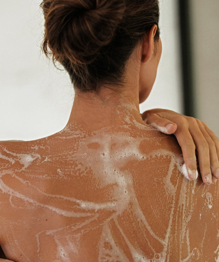 Seen from behind, a person with their hair in a bun cleanses their upper back with Awaken Body Wash, creating suds and bubbles as they reach over the shoulder to wash, enjoying the scent of Lavender and Cedar Oil.