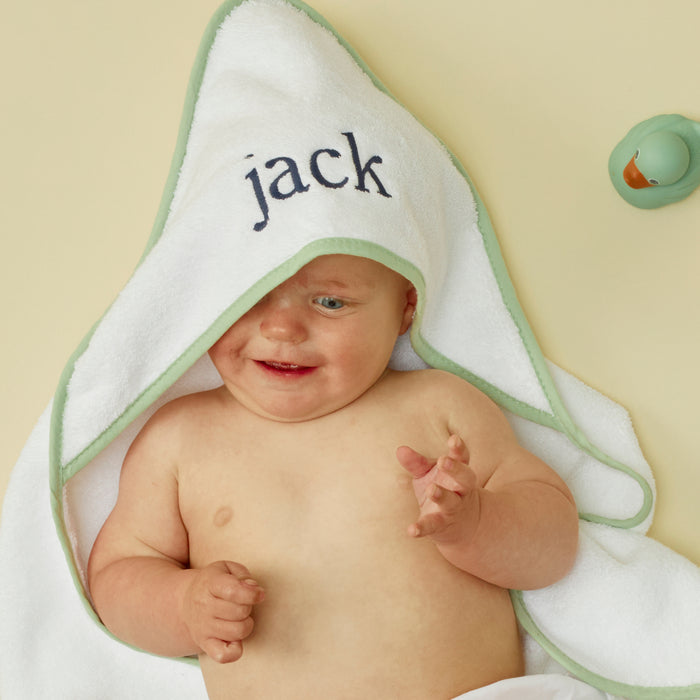 Smiling baby wearing a white hooded towel with matcha green piping and "jack" embroidered on the hood.
