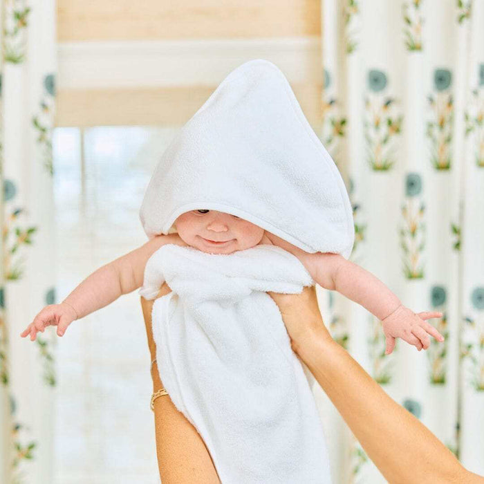 A baby wrapped in a soft white hooded towel, held up by an adult's hands in front of patterned curtains.