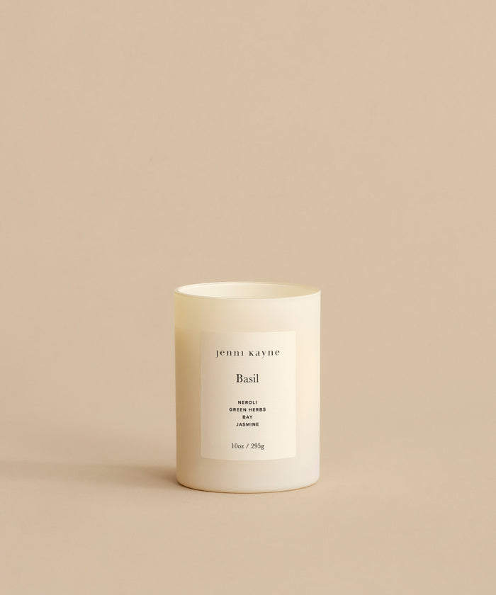 A white Basil Glass Candle labeled jenni kayne sits on a beige surface. Hand-poured in Los Angeles, it features notes of neroli, green herbs, and jasmine—a blend from Oprah’s Favorite Things.