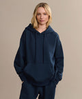 A blonde woman wears a loose-fitting navy Bay Hoodie with matching pants, standing with hands in the pocket against a plain beige background.