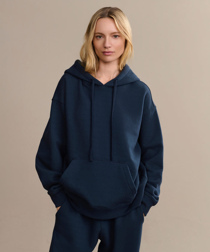 A blonde woman wears a loose-fitting navy Bay Hoodie with matching pants, standing with hands in the pocket against a plain beige background.
