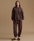 A woman stands against a plain warm beige studio background wearing a matching dark brown hoodie and jogger sweatpants set, with brown slip-on slippers.