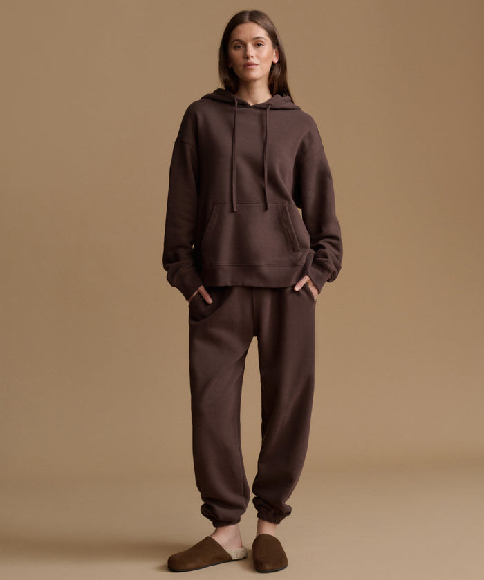 A woman stands against a plain warm beige studio background wearing a matching dark brown hoodie and jogger sweatpants set, with brown slip-on slippers.