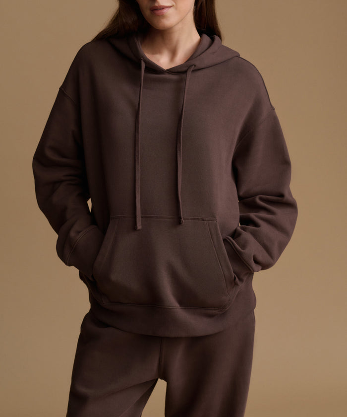A model wearing a dark brown oversized pullover hoodie with matching sweatpants, shown from the neck down against a solid warm beige studio background.