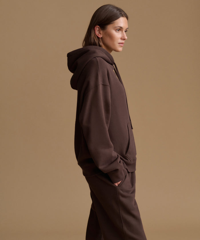 A woman in side profile wearing a dark brown hooded sweatshirt and matching sweatpants against a solid warm beige studio background.