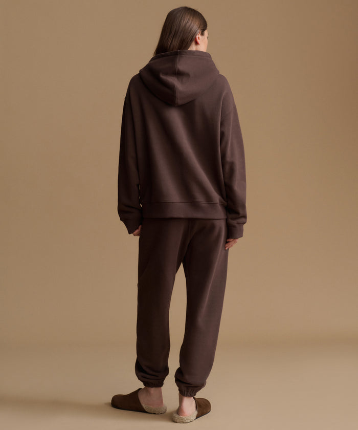 A model viewed from behind wearing a matching dark brown hoodie and jogger sweatpants set, standing against a solid beige studio background.