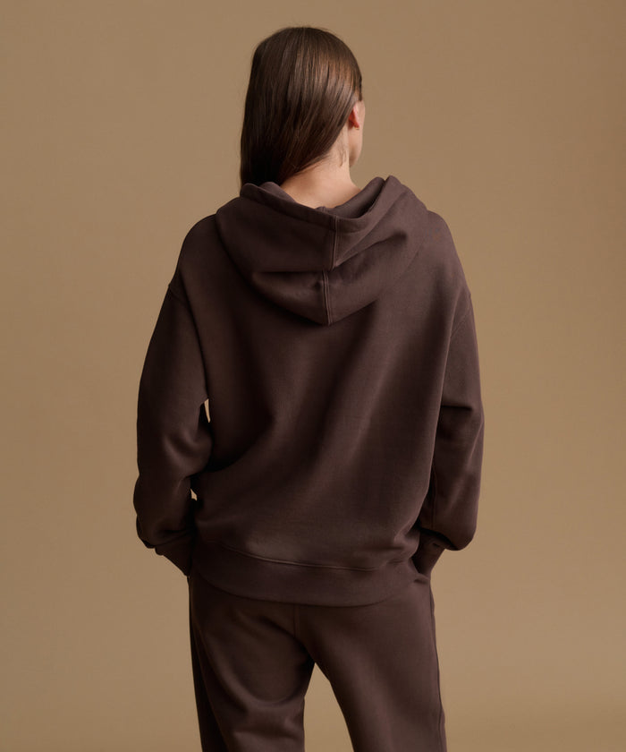 A model shown from the back wearing a dark brown oversized hoodie and matching dark brown sweatpants against a solid warm beige studio background.