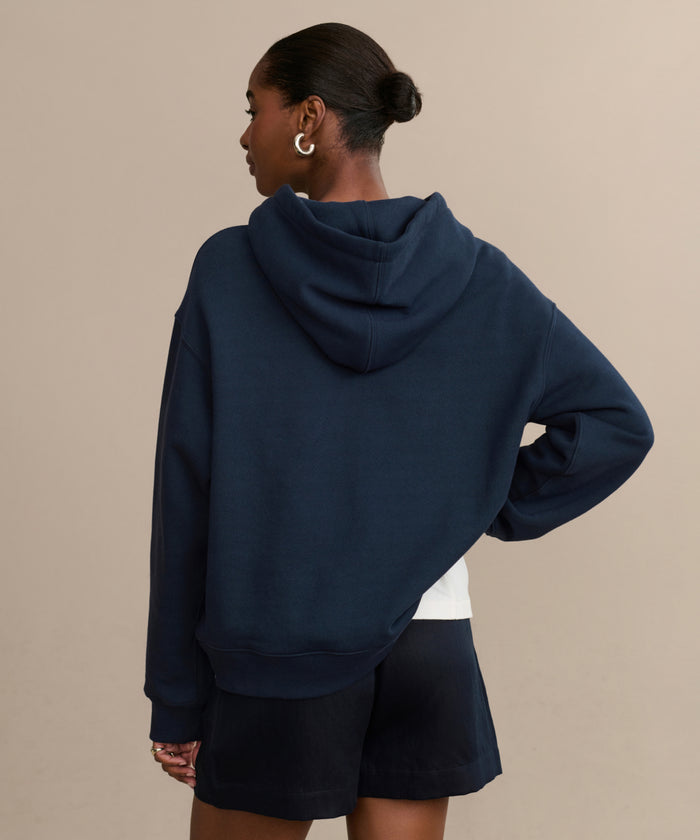 A person with short hair in a bun is seen from behind, wearing the Bay Hoodie in navy brushed fleece with matching shorts and one hand on their hip, set against a plain beige background.