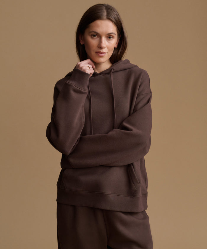 A woman modeling a dark brown pullover hoodie and matching sweatpants against a solid warm beige studio background.