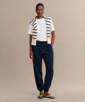 Against a beige background, a woman wears a white T-shirt, navy Bay Sweatpant joggers in brushed fleece, sandals, and has a white sweater with navy stripes draped over her shoulders, with her hands in her pockets.
