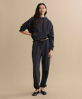 A woman with long dark hair poses casually against a beige background, wearing a black hoodie and the Bay Sweatpant in athletic brushed fleece, paired with black flats.