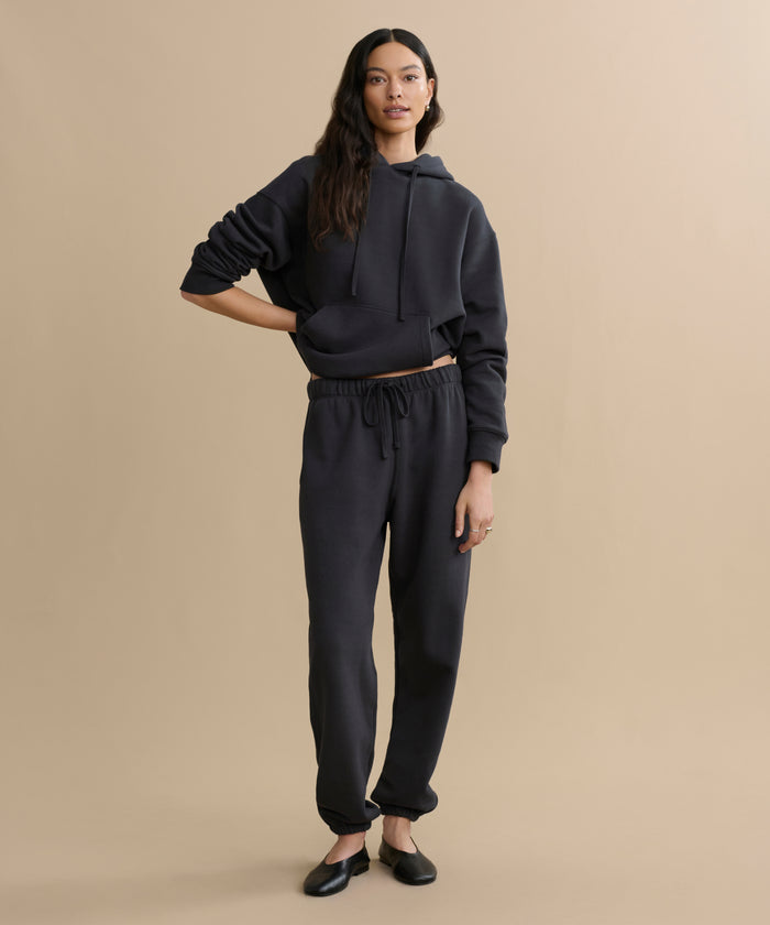 A woman with long dark hair poses casually against a beige background, wearing a black hoodie and the Bay Sweatpant in athletic brushed fleece, paired with black flats.