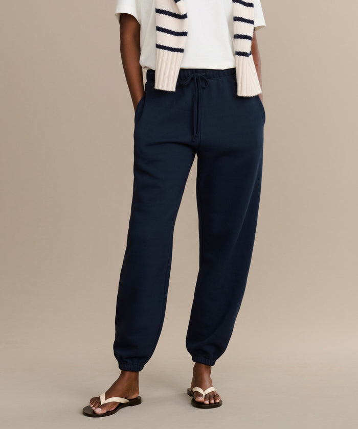 Someone wearing navy blue Bay Sweatpants, a white t-shirt, a cream and navy striped sweater over their shoulders, and beige sandals stands against a plain beige background.