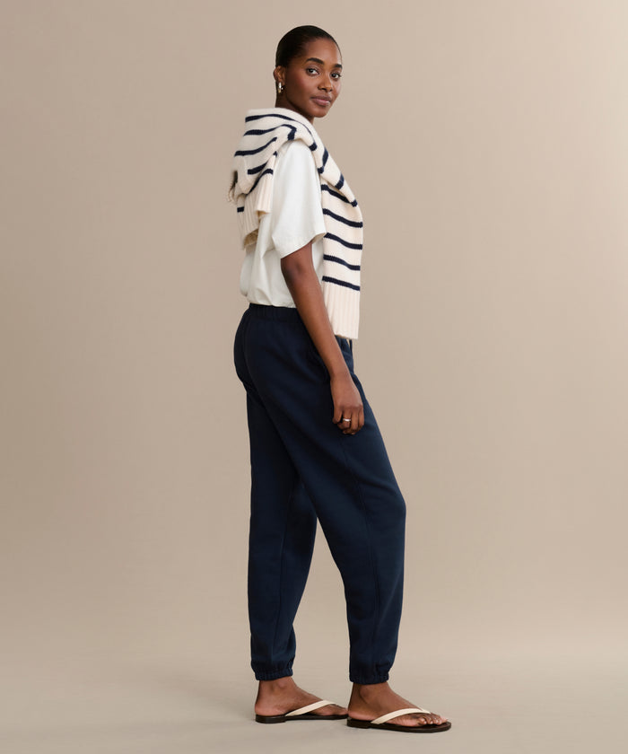 A woman stands sideways wearing a white shirt, navy Bay Sweatpant joggers, sandals, and a cream sweater with navy stripes draped over her shoulders. The background is plain beige.