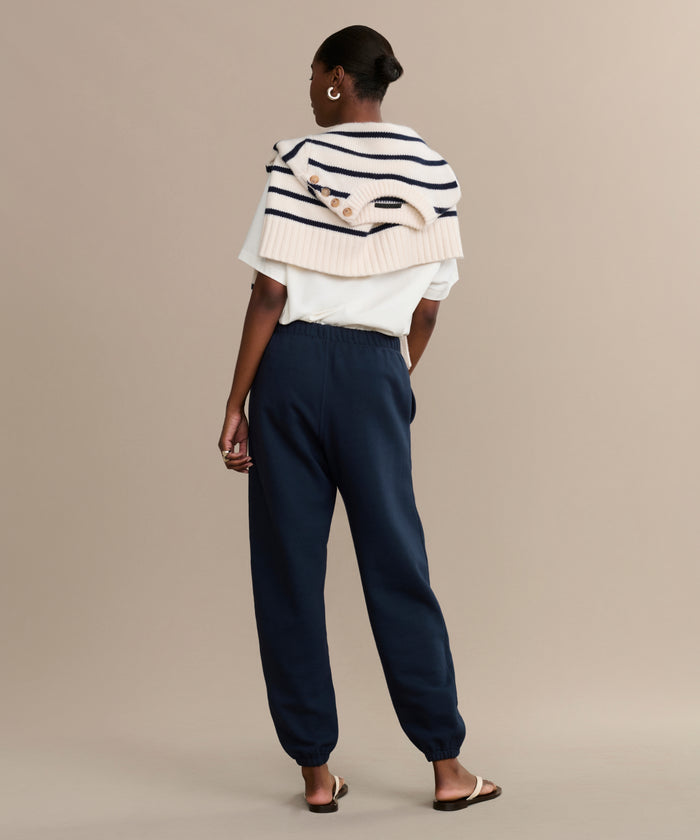A woman with neatly styled short hair stands facing away in a white T-shirt, Bay Sweatpant athletic sweats, and sandals. A striped sweater is draped over her shoulders against a plain beige background.