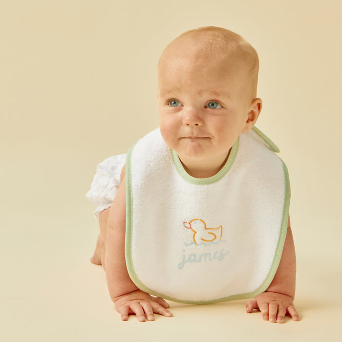 Crawling baby in a white bib with matcha green trim, duck embroidery, and James.