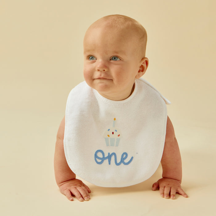 Baby wearing a white first birthday bib with blue 'one' and cupcake.