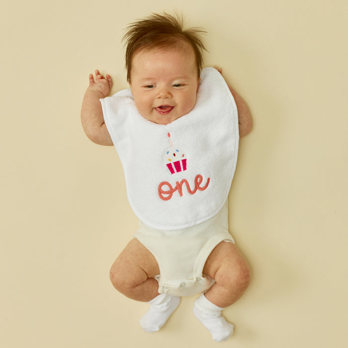 Baby wearing a white first birthday bib with pink 'one' and cupcake.