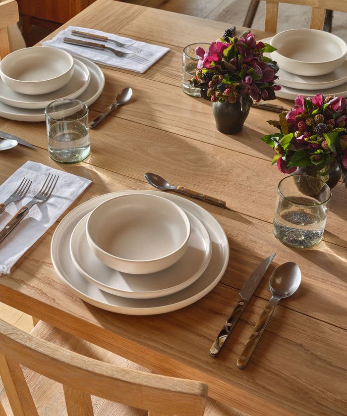 A wooden dining table set with white plates, bowls, a Bistro Vintage Finish Cutlery Set, drinking glasses, and white napkins. Two small vases with purple and green flowers serve as centerpieces in the sunlight.