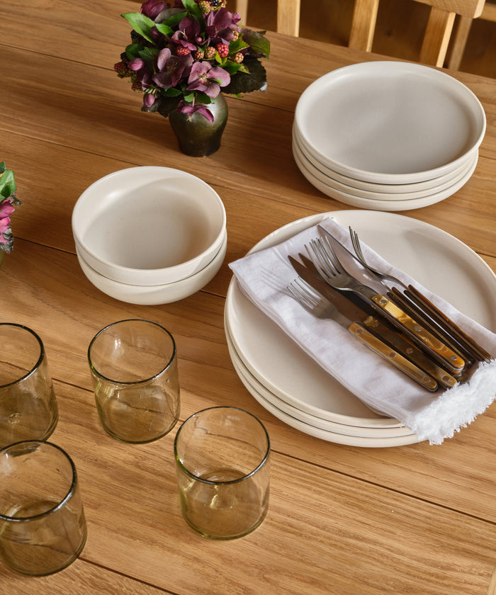 A wooden table set with stacked white plates, bowls, and glasses features the Bistro Vintage Finish Cutlery Set with brown handles on a white napkin, accented by small flower arrangements as decoration.