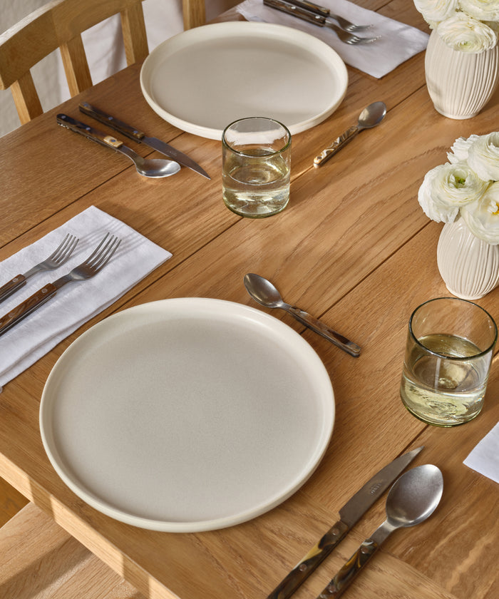A wooden dining table set for two features white plates, glasses of water, and the Bistro Vintage Finish Cutlery Set on white napkins. White vases with white flowers enhance the charm of this simple, neatly arranged setting.