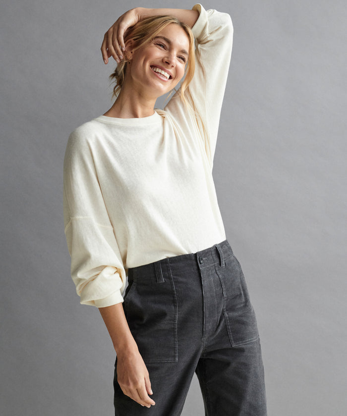 A smiling blonde woman wears the Boyfriend Long-Sleeve Tee with high-waisted dark pants, posing with one arm bent behind her head against a plain gray background.