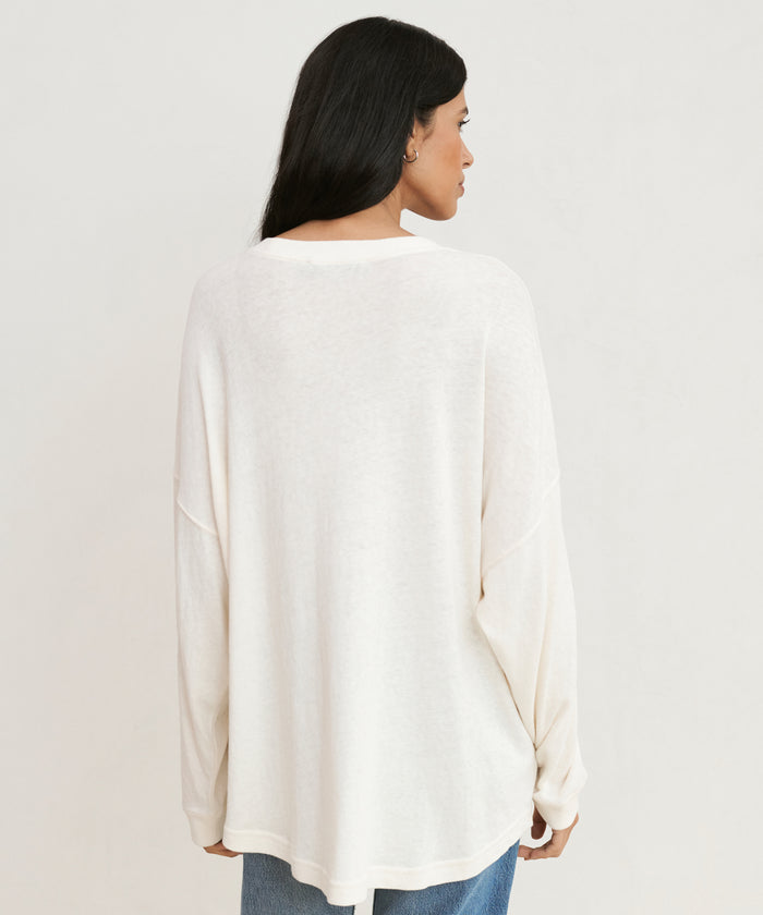 A woman with long dark hair stands with her back to the camera, dressed in an off-white Boyfriend Long-Sleeve Tee and blue jeans, styled casually against a plain light background.