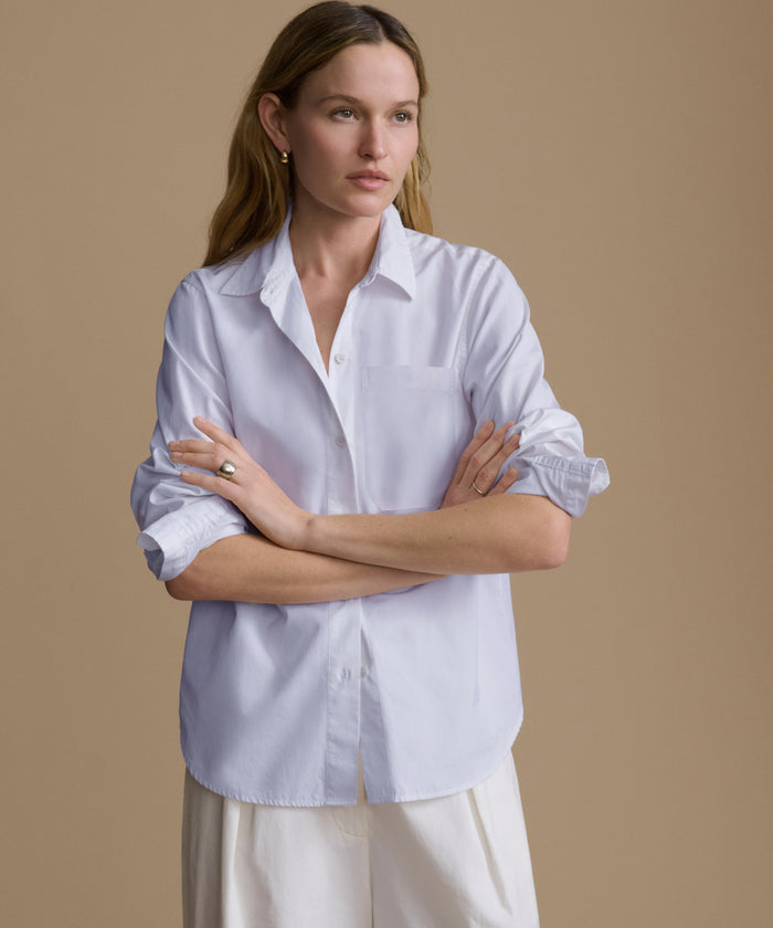 A woman modeling a light blue long-sleeve button-up shirt with white trousers, posed against a solid beige studio background.
