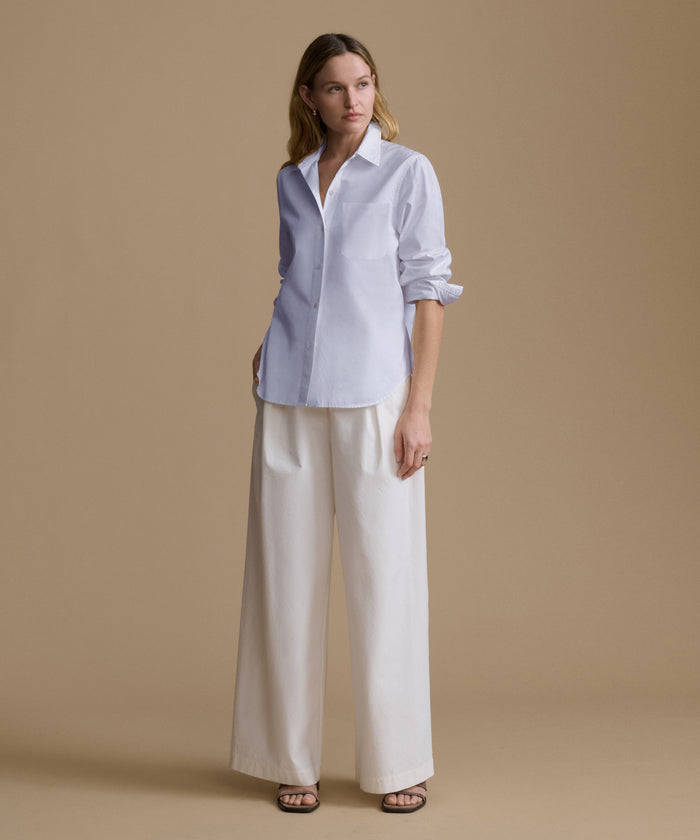 A woman stands against a plain beige studio background wearing a light blue button-up shirt with rolled sleeves and wide-leg white pants.