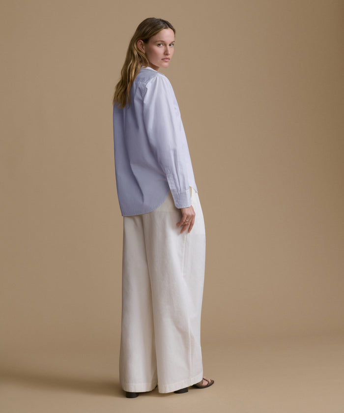 A woman stands in profile wearing a light blue long-sleeve blouse with wide-leg off-white trousers, photographed against a solid warm beige studio background.