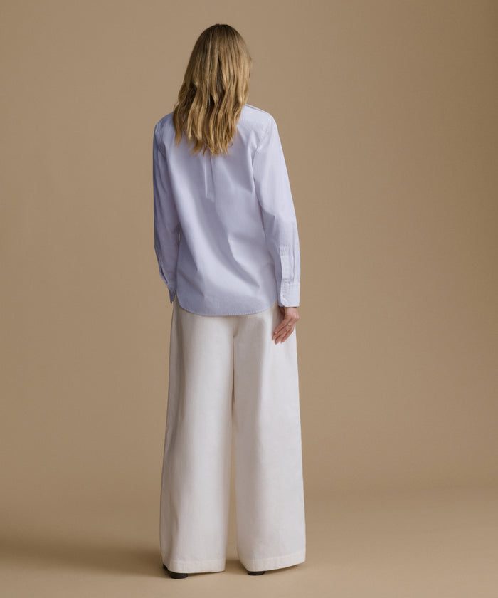 A woman viewed from behind wearing a light blue long-sleeve button-up shirt and wide-leg white trousers, standing against a solid beige studio background.
