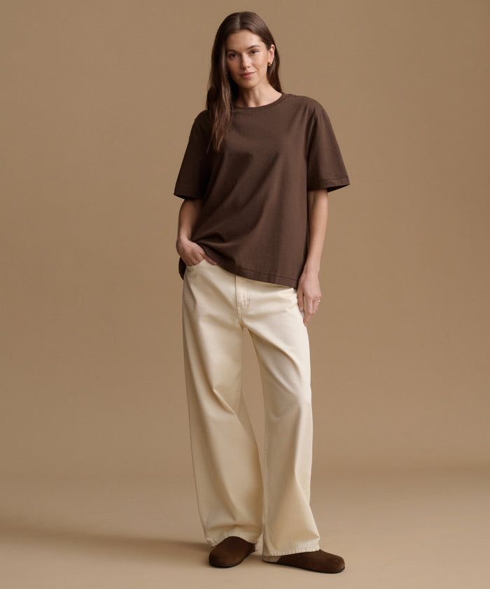 A woman modeling an oversized brown short-sleeve t-shirt and cream wide-leg pants against a solid beige studio background.