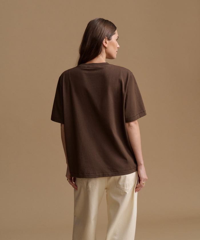 A woman shown from the back wearing an oversized dark brown short-sleeve t-shirt and cream-colored pants, standing against a solid beige studio background.