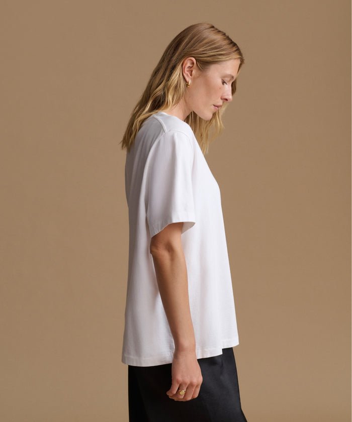 A woman in profile wearing a loose white short-sleeve t-shirt and black pants, standing against a solid beige studio background.