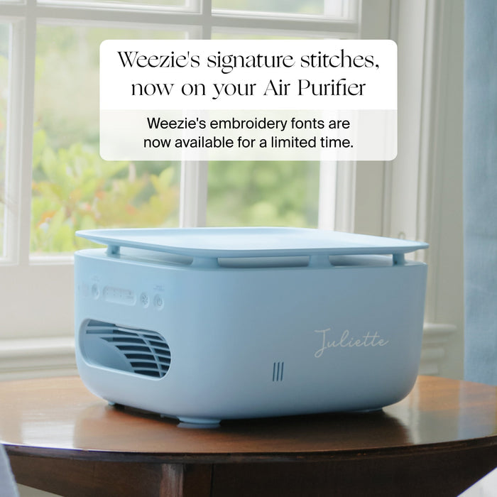A light blue Air Purifier placed on a wooden table in front of a window, with a text overlay that reads 'Weezie's signature stitches, now on your Air Purifier' and 'Weezie's embroidery fonts are now available for a limited time.' The word 'Juliette' is embroidered on the side of the device.