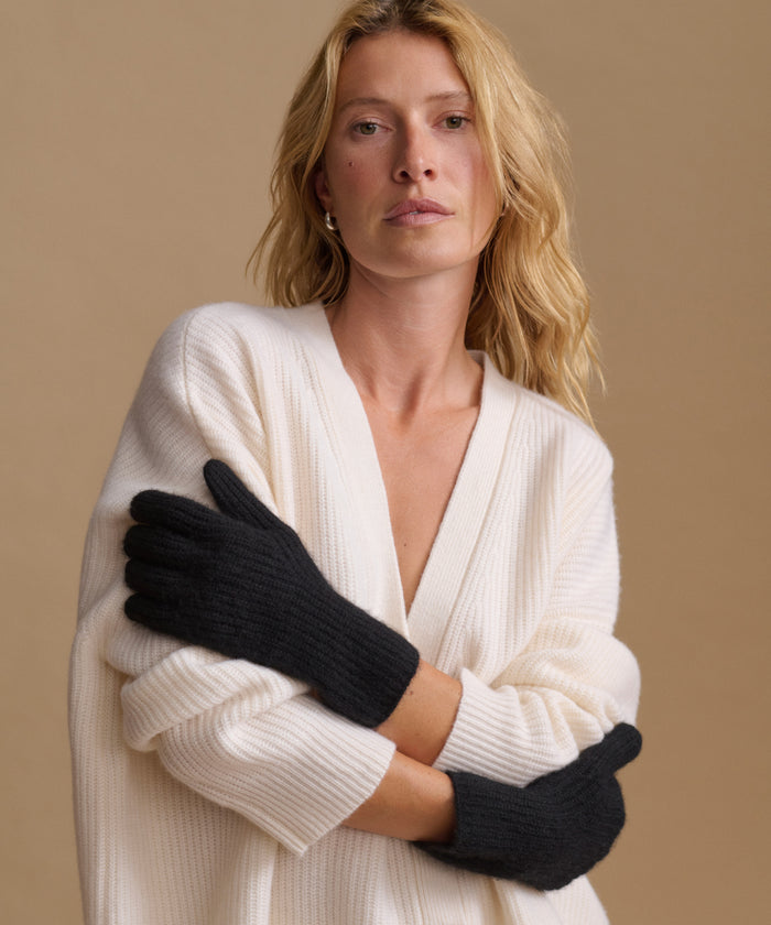 A woman wearing black knit gloves and a cream ribbed cardigan against a solid beige background.