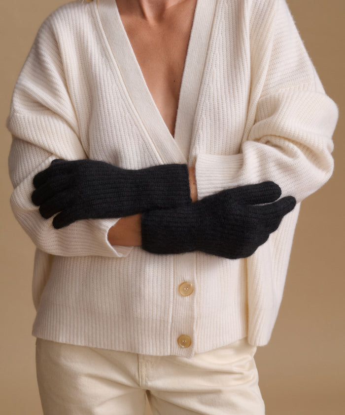 A person wearing a cream ribbed cardigan and cream pants, with black knit gloves crossed over the chest, against a warm beige studio background.